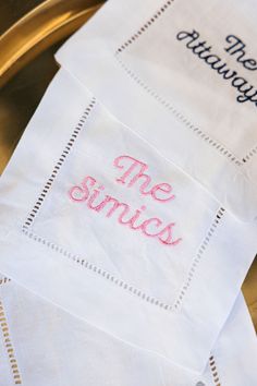 three napkins with embroidered words on them sitting on a gold tray next to a cup and saucer