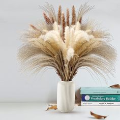 a white vase filled with dry grass next to two books