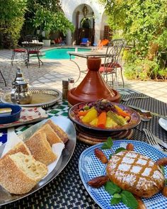 an outdoor table with plates and bowls of food on it, near a swimming pool