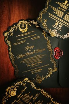 two black and gold wedding cards with red wax stamp on them sitting on a wooden table