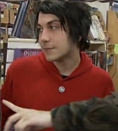 a young man with black hair is talking to someone in front of bookshelves