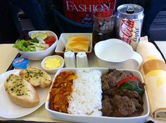 a tray with rice, meat and vegetables on it next to two sodas as well as other food items