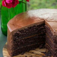 a chocolate cake with one slice missing from it on a wooden platter next to a green vase filled with flowers