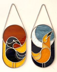 two stained glass birds hanging from chains on a white wall, one is orange and the other is blue