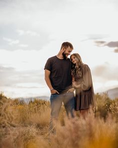 a man and woman are standing in tall grass with their arms around each other as the sun sets behind them