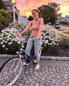 a woman standing next to a bike on a sidewalk with flowers in the back ground