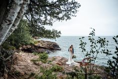 two brides standing on the edge of a cliff looking out at the water and trees