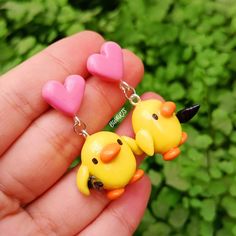 two small yellow birds with pink hearts hanging from them's earring hooks in front of some green plants