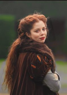 a woman with long red hair wearing a brown cape and holding her hand on her shoulder