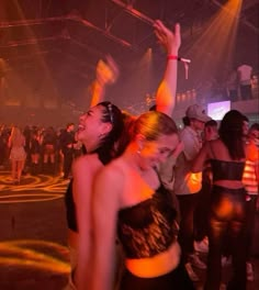 two women dancing at a party with other people in the back ground and lights on