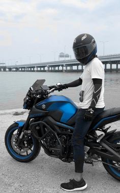 a person sitting on a blue motorcycle near the water with a bridge in the background