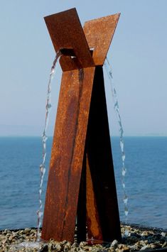 a metal sculpture sitting on top of a rock covered beach next to the ocean with water coming out of it