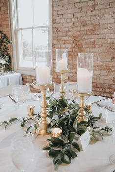 a table with candles and greenery on it in front of a brick wall at a wedding reception