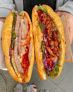 two sandwiches with meat and vegetables in buns held by someone's hands on the sidewalk