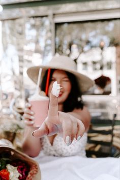 a woman in a white dress and hat pointing at something with her hand on the table