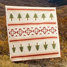 a white quilt with trees on it sitting in the grass near some bushes and mountains