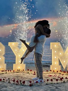 a man holding a woman in his arms on the beach with fireworks behind him and letters spelling love