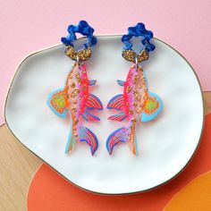 two colorful fish shaped earrings on a white plate next to an orange and pink background