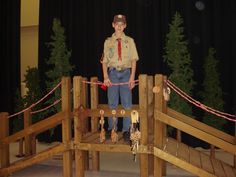 a boy in scout uniform standing on a bridge made out of wooden planks and ropes