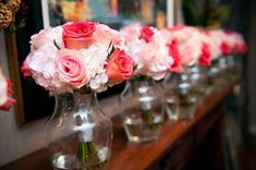 pink and white flowers in vases lined up on a long wooden table with framed pictures behind them