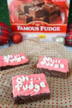 some brownies with pink frosting on top of a table next to a box of famous fudge