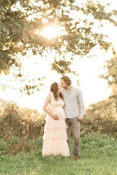 a pregnant woman standing next to a man in a field with the sun behind her