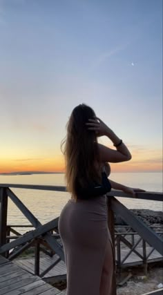 a woman standing on top of a wooden pier next to the ocean at sunset with her hair blowing in the wind