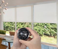 a person holding a clock in front of a dining room window with white roller shades