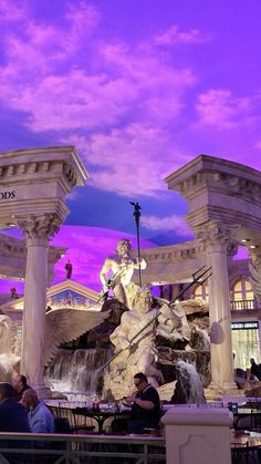 people sitting at tables in front of a fountain with statues on it and purple skies above