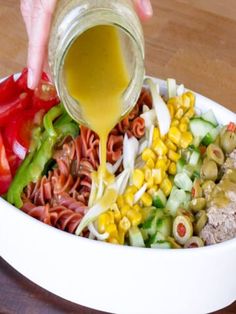 a person pouring dressing into a bowl filled with vegetables and pasta salads on a wooden table