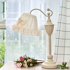 a table with a lamp and flowers on it next to a window sill in front of a curtain