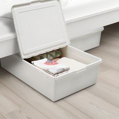 an open storage box sitting on the floor in front of a bed with white sheets and pillows