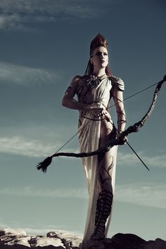 a woman dressed in white holding a bow and arrow on top of a rocky hill