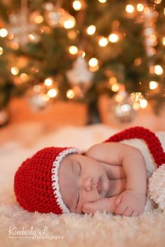 a baby is wearing a red and white crocheted santa hat while laying in front of a christmas tree