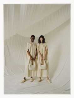 two women standing in front of a white backdrop wearing capes and holding purses