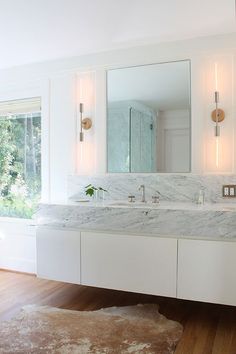 a white bathroom with marble counter tops and double sinks, along with two mirrors on the wall