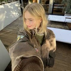 a woman with blonde hair wearing a brown jacket and looking at the camera while standing on a wooden deck