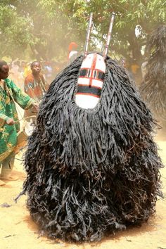 a man standing next to a large black object on top of dirt covered ground with trees in the background