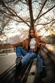 a woman sitting on top of a wooden bench next to a leaf filled tree in a park