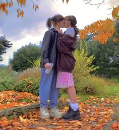 two people standing in front of a tree with leaves on the ground and one person kissing the other