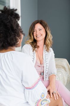 a woman sitting on a couch talking to another woman who is wearing pink pants and a white shirt