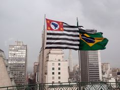 two flags flying next to each other in front of tall buildings on a cloudy day