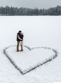 a man and woman standing in the middle of a heart shaped snow covered field with their arms around each other