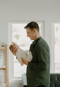 a man holding a baby in his arms while standing next to a couch and window