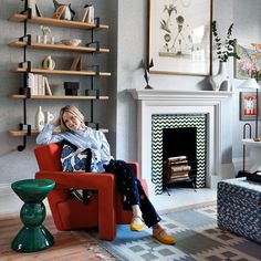 a woman sitting in a red chair next to a fire place and bookshelf