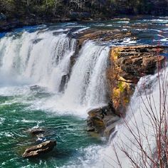 the waterfall is surrounded by green water and brown rocks with trees in the background, while two boats are on the riverbank