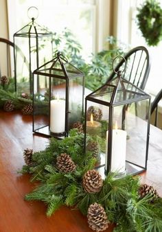 three glass lanterns sitting on top of a wooden table next to pine cones and greenery