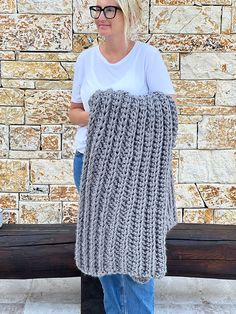 a woman standing next to a wooden bench holding a gray knitted blanket in front of a brick wall