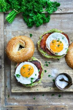 three bagels with eggs and bacon on them are sitting on a wooden cutting board