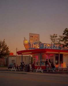 people are sitting at tables outside of a frozen food stand with neon signs on the roof